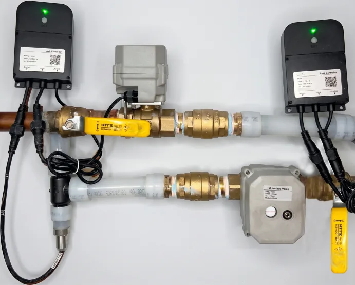 An in-suite installation on hot and cold lines: two leak controllers, motorized shutoff valves on the ball valves, brass unions, and an in-line flow sensor probe fitted into the cold supply.